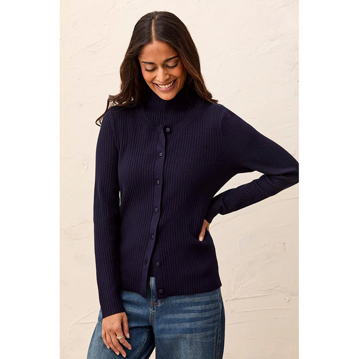 A woman wearing a dark navy ribbed button-front sweater and blue jeans, standing against a light beige textured wall.