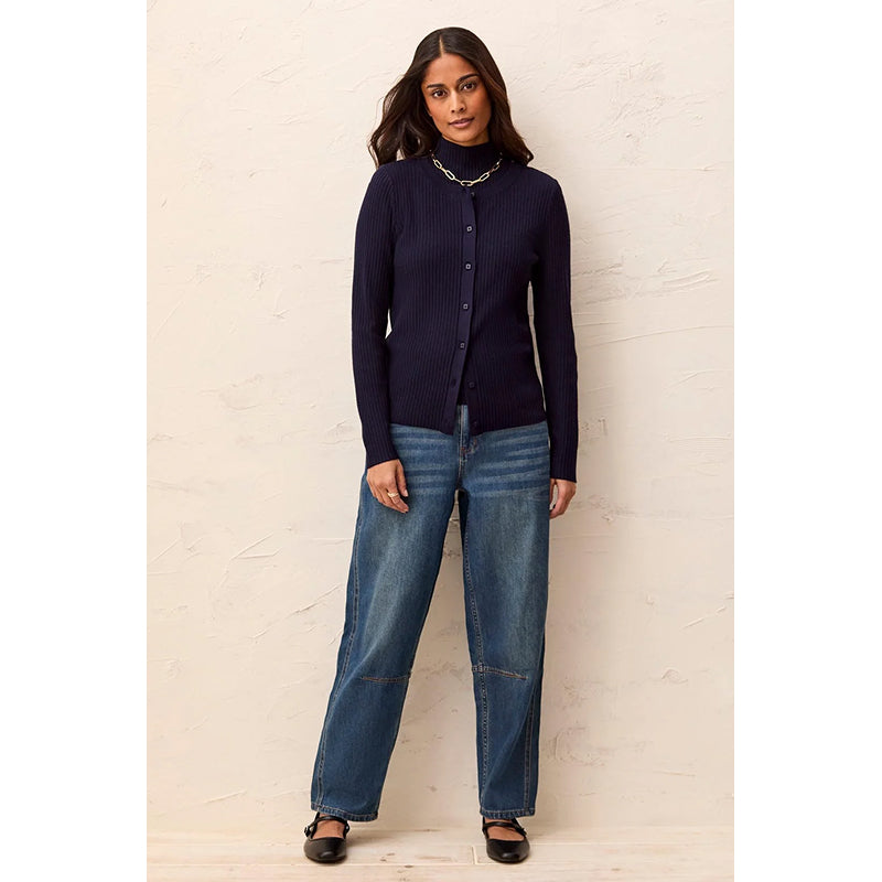 A woman modeling a dark navy ribbed button-up cardigan with blue jeans and black flat shoes, standing against a textured off-white wall.