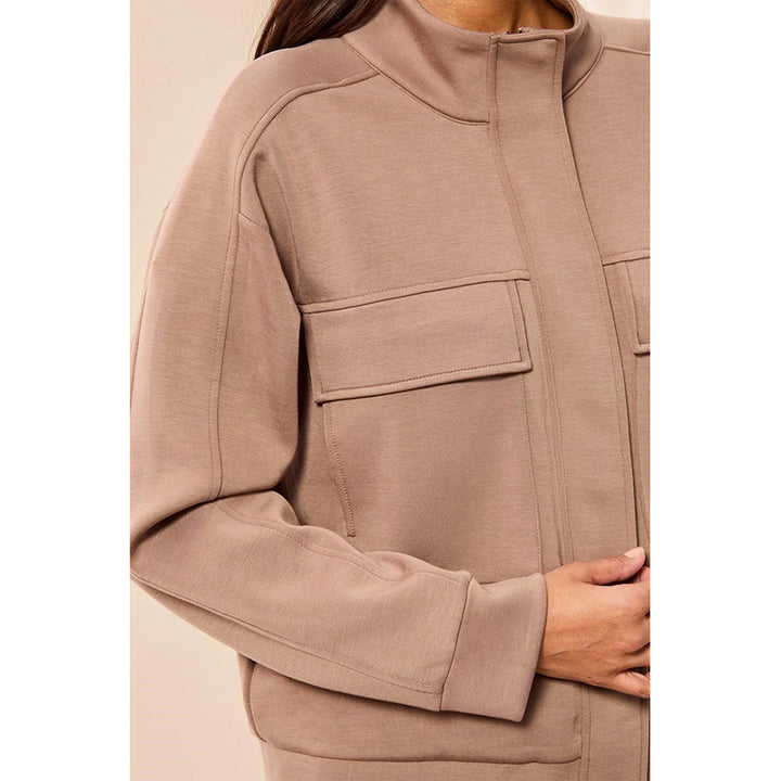 A close-up of a person wearing a taupe-brown zip-up jacket with a mock neck and flap chest pockets, shown against a plain beige background.