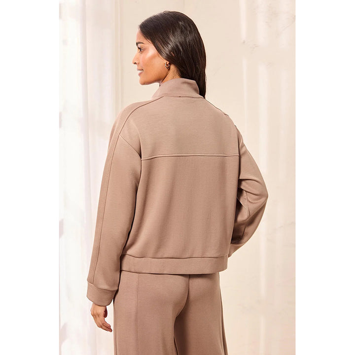 A woman shown from the back wearing a taupe-brown cropped jacket and matching pants against a soft cream indoor backdrop.