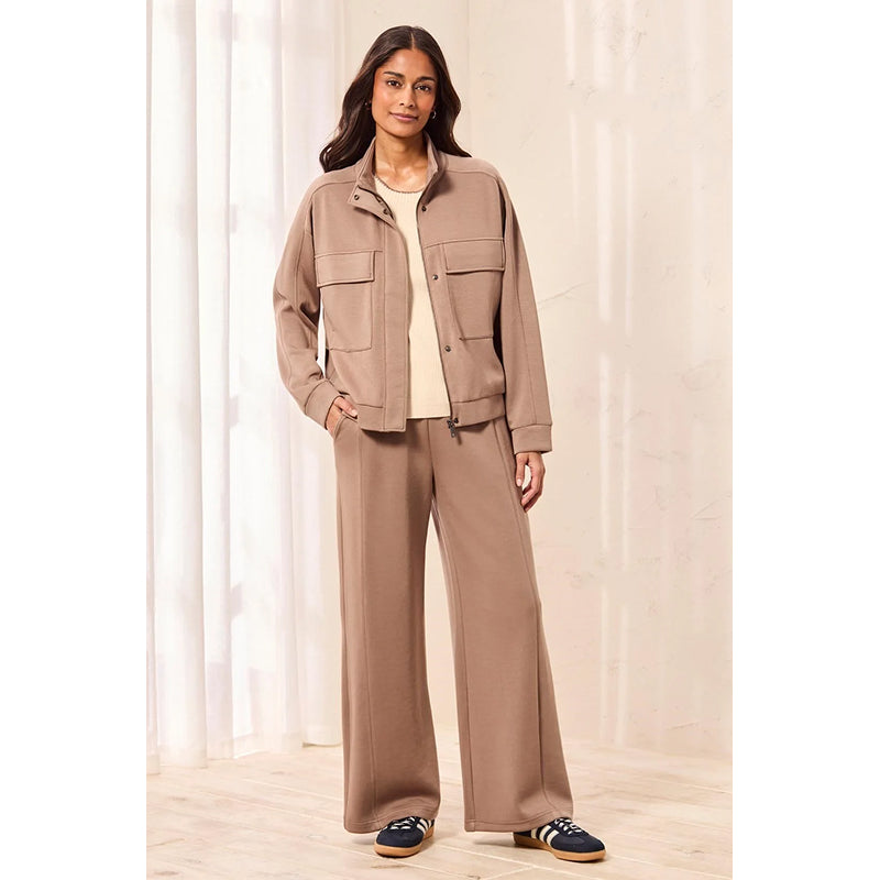 A woman modeling a matching taupe-brown two-piece set consisting of a cropped utility jacket with flap pockets and wide-leg trousers, styled over a cream t-shirt and navy sneakers.
