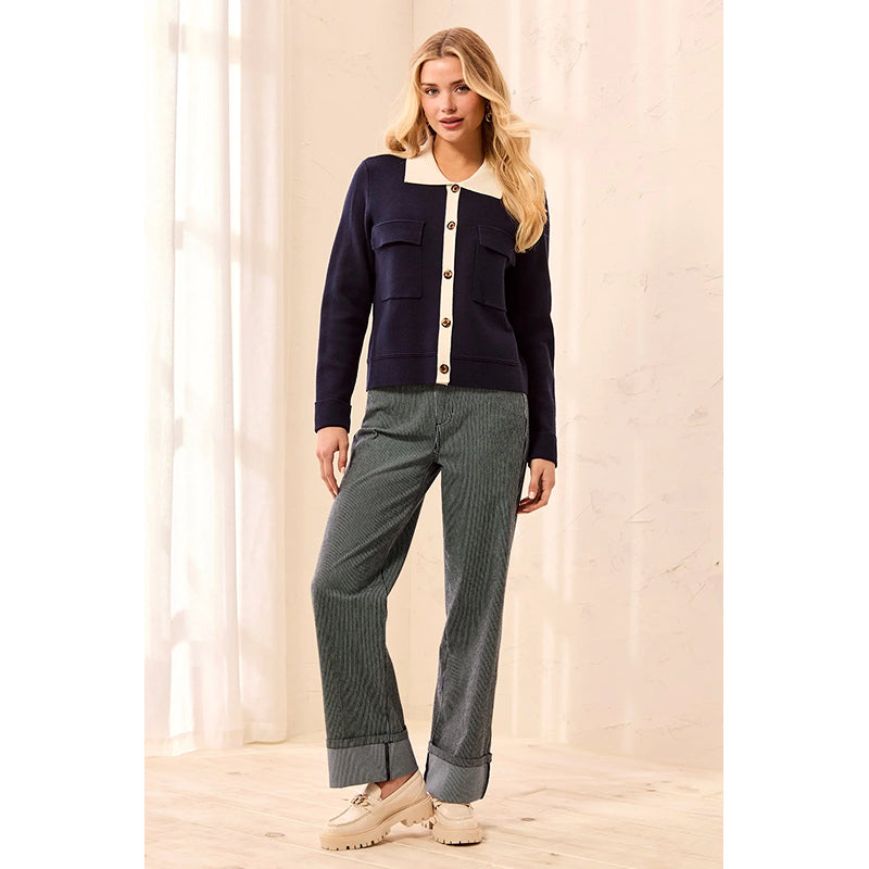 A woman models a navy blue knit cardigan with a cream collar and button placket, paired with gray pinstriped trousers and beige platform loafers, set against a warm beige background with soft window light.