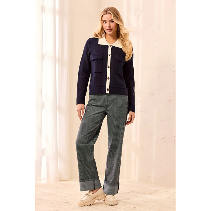 A woman models a navy blue knit cardigan with a cream collar and button placket, paired with gray pinstriped trousers and beige platform loafers, set against a warm beige background with soft window light.