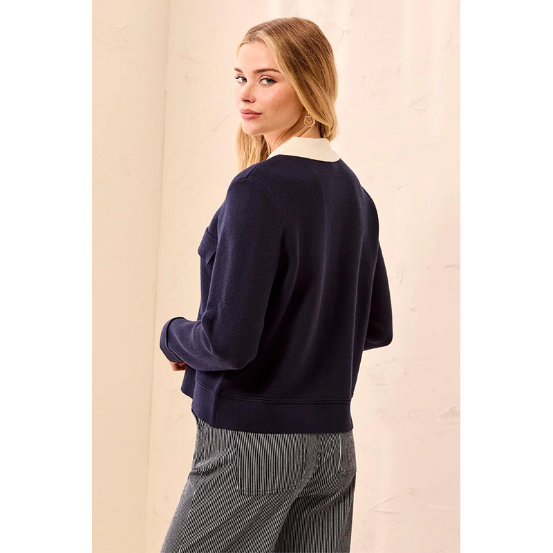 A woman wearing a navy blue knit sweater with a cream ribbed collar, shown from the back against a light beige textured wall.