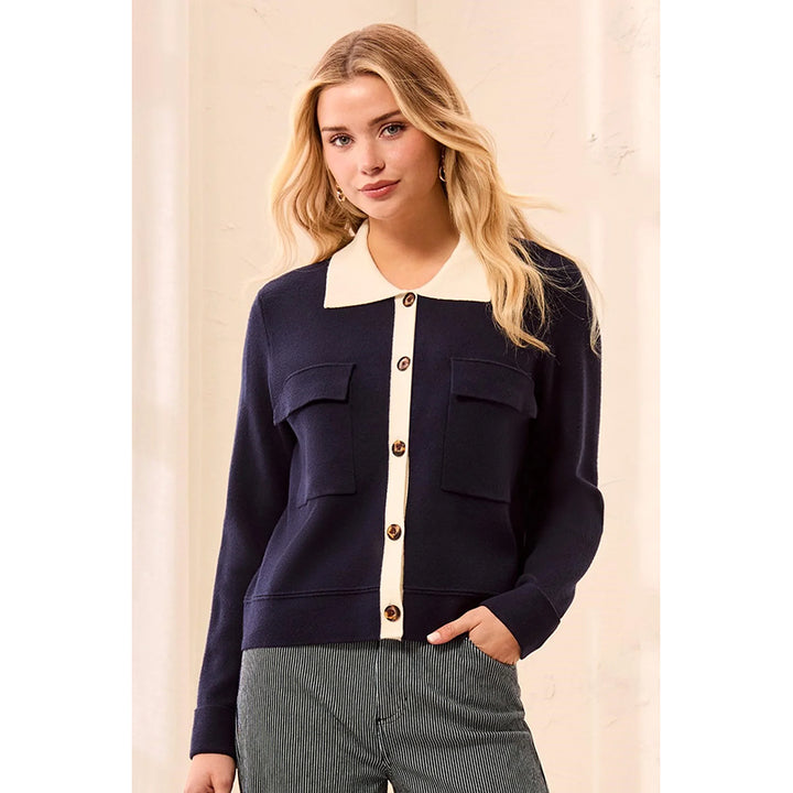 A woman wearing a navy blue knit cardigan with a cream-colored collar and button placket, paired with gray textured pants, set against a neutral cream-colored background.