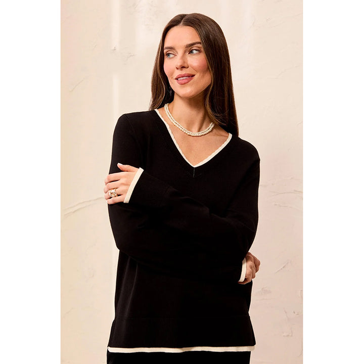A woman modeling a black long-sleeve knit sweater with contrasting white trim along the V-neck, cuffs, and hem, set against a light beige textured wall.