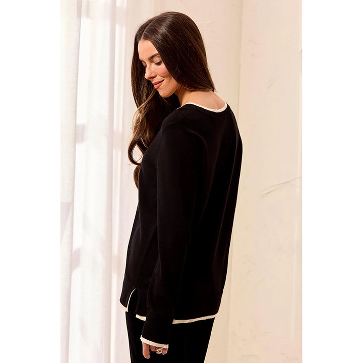 A woman wearing a black long-sleeve top with white contrast trim, shown from the back in a softly lit indoor setting with sheer curtains.