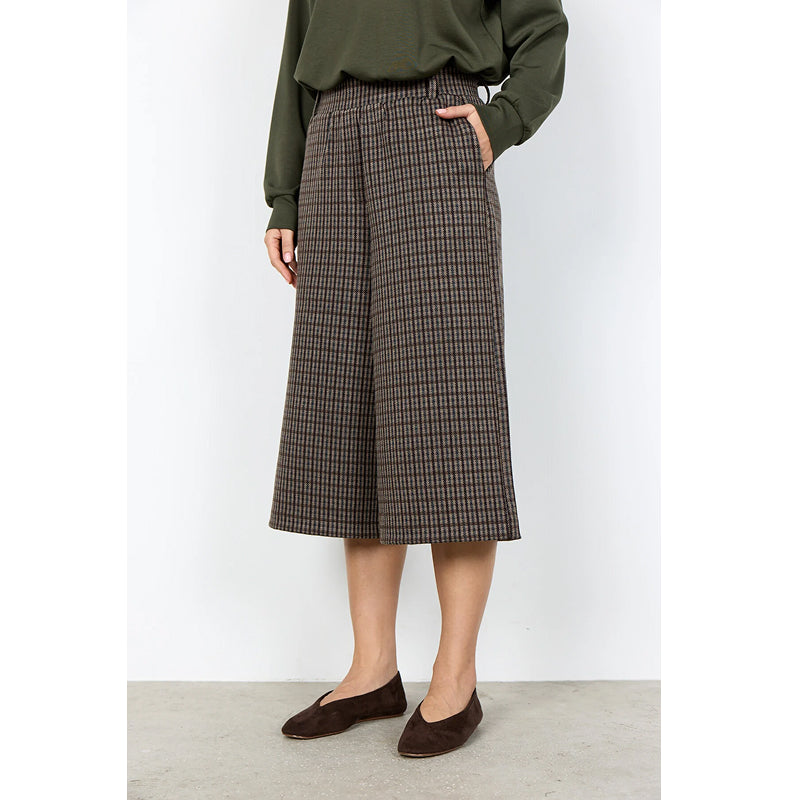 A model wearing a brown and beige houndstooth midi skirt with a wide leg, paired with an olive green long-sleeve top and dark brown suede flats, standing against a white wall.