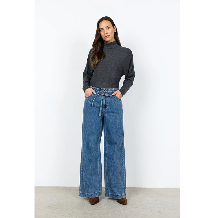 A woman modeling a dark gray mock neck knit top and high-waisted, wide-leg blue denim jeans against a white studio background.