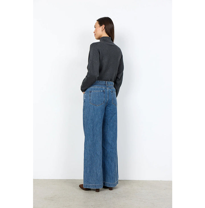 A woman stands with her back to the camera wearing a dark charcoal gray mock-neck knit top and high-waisted, wide-leg blue denim jeans, set against a minimalist white wall and light concrete floor.