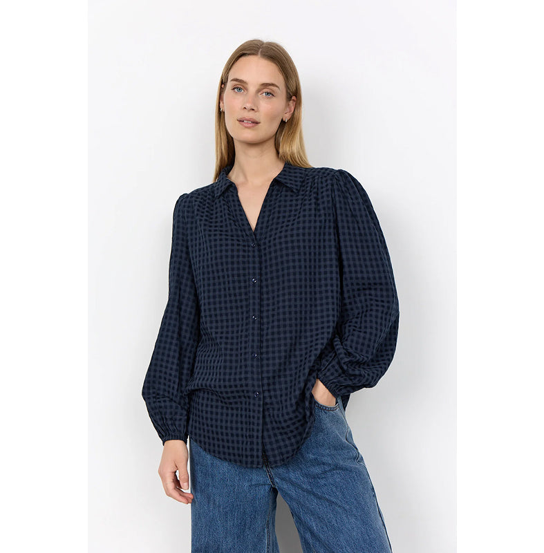 A woman stands against a plain white wall wearing a dark navy and black gingham long-sleeve blouse with puffed shoulders and wide-leg blue jeans.