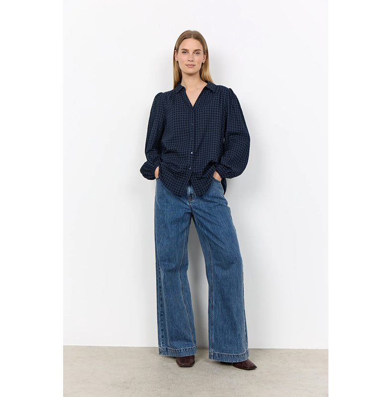 A woman stands against a plain white wall wearing a dark navy and black gingham long-sleeve blouse with puffed shoulders and wide-leg blue jeans.