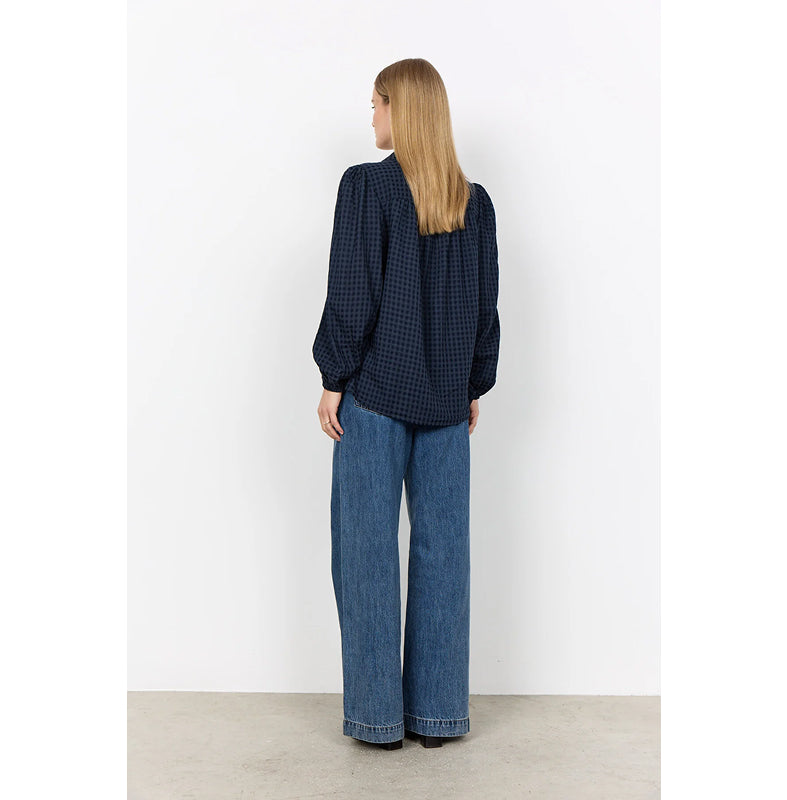 A woman stands with her back to the camera wearing a navy blue and black gingham long-sleeve blouse with puffed shoulders, paired with wide-leg blue jeans, in front of a white wall.
