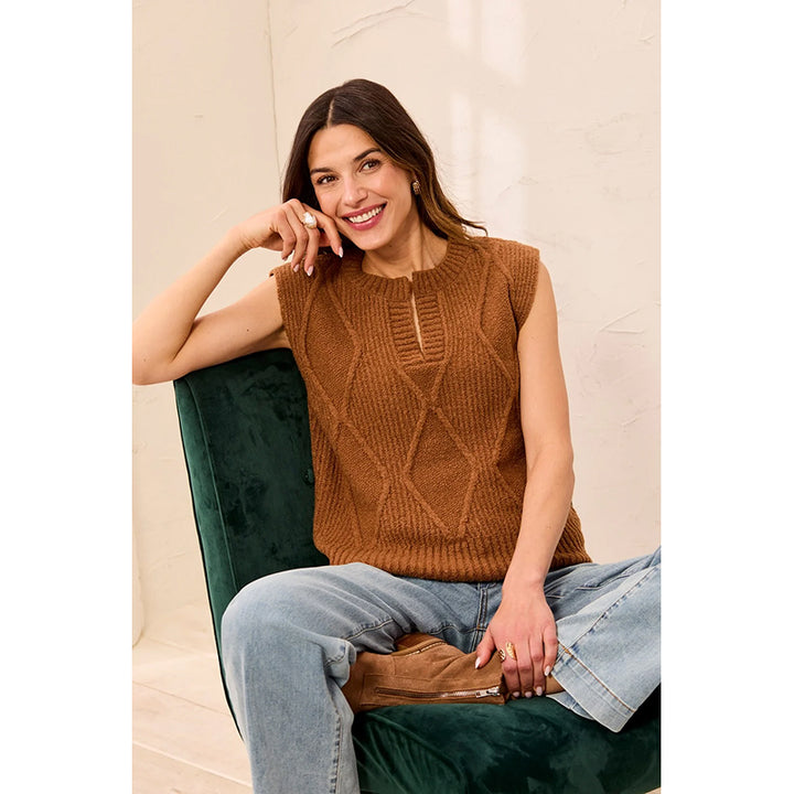 A woman wearing a sleeveless brown cable-knit sweater vest with a split neckline sits on a dark green velvet chair.