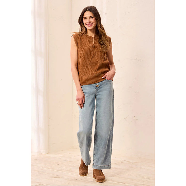 A woman modeling a sleeveless brown cable-knit sweater vest paired with light-wash wide-leg jeans and brown suede boots, standing in a bright, neutral indoor setting.