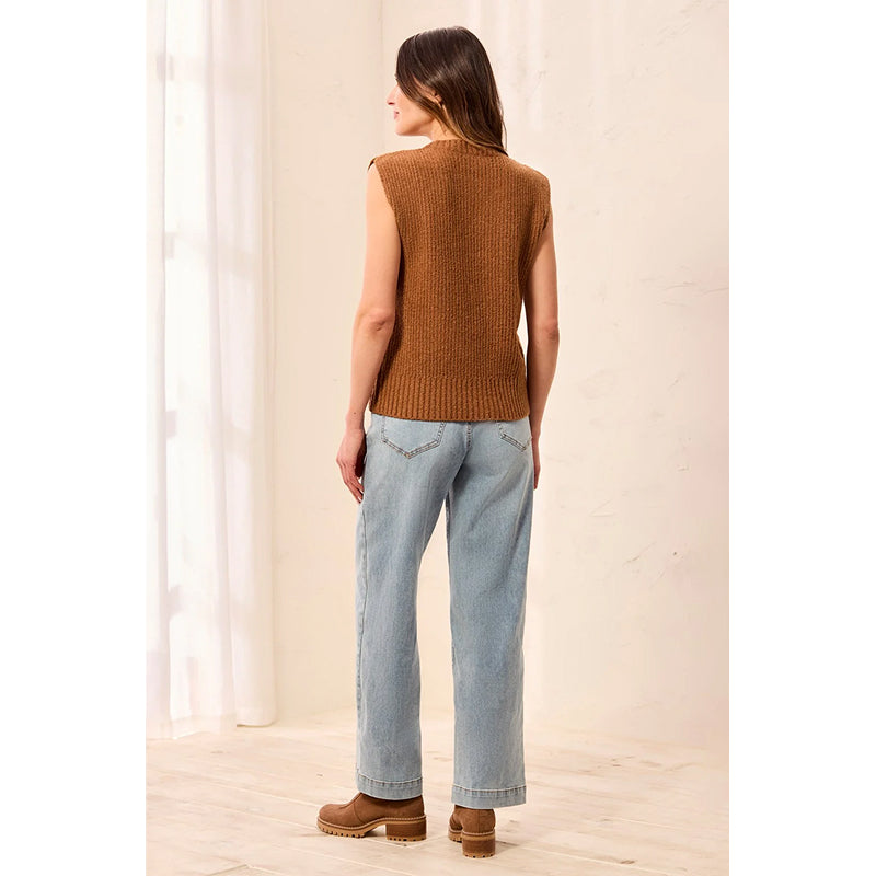 A woman is shown from behind wearing a sleeveless brown ribbed knit vest, light-wash blue jeans, and brown platform boots in a bright, minimalist indoor setting.