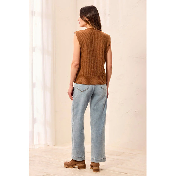 A woman is shown from behind wearing a sleeveless brown ribbed knit vest, light-wash blue jeans, and brown platform boots in a bright, minimalist indoor setting.