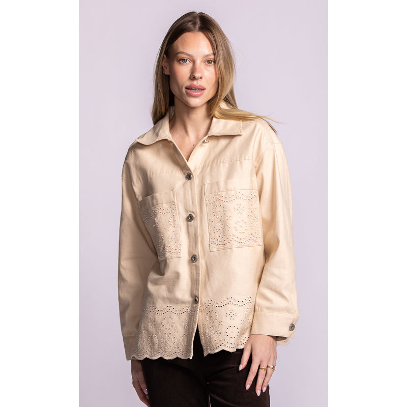 A woman modeling a light beige long-sleeve button-up shirt with embroidered and eyelet lace details on the chest and lower front, paired with dark pants against a solid light lavender background.