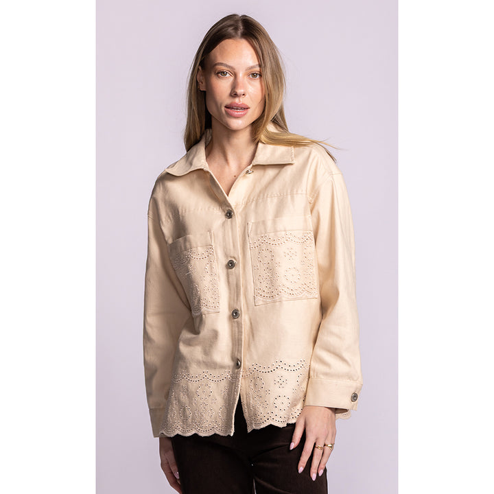 A woman modeling a light beige long-sleeve button-up shirt with embroidered and eyelet lace details on the chest and lower front, paired with dark pants against a solid light lavender background.