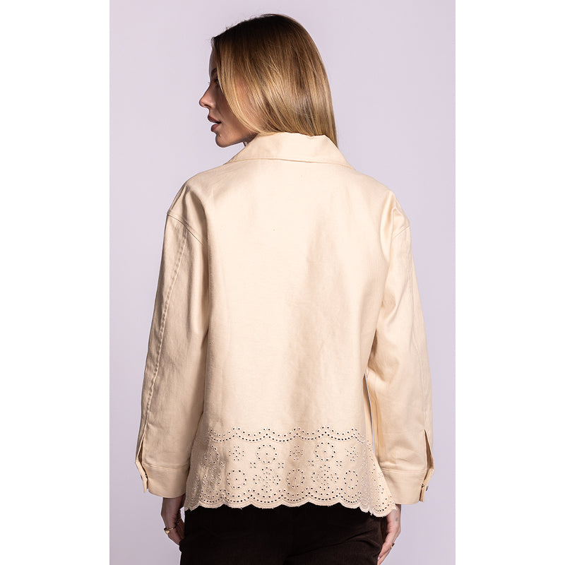 A woman shown from the back wearing a light beige long-sleeve jacket with a scalloped eyelet lace hem, paired with dark brown pants, against a solid light lavender background.
