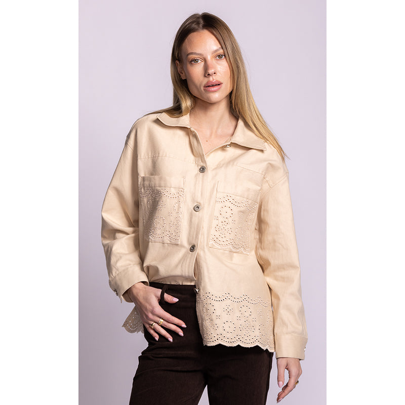 A woman modeling a light beige button-up shirt with eyelet lace details on the chest and hem, paired with dark brown pants, against a solid light purple background.