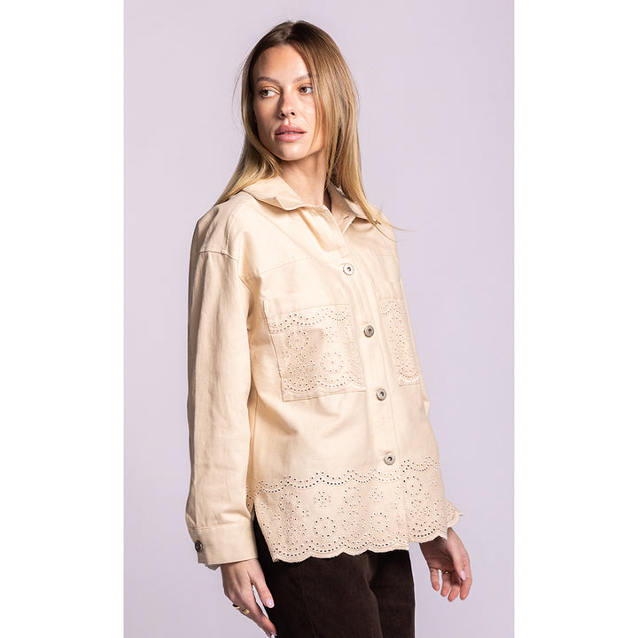 A woman modeling a light beige long-sleeve button-up shirt with eyelet lace detailing on the chest pockets and hem, paired with dark brown pants against a solid light lavender background.