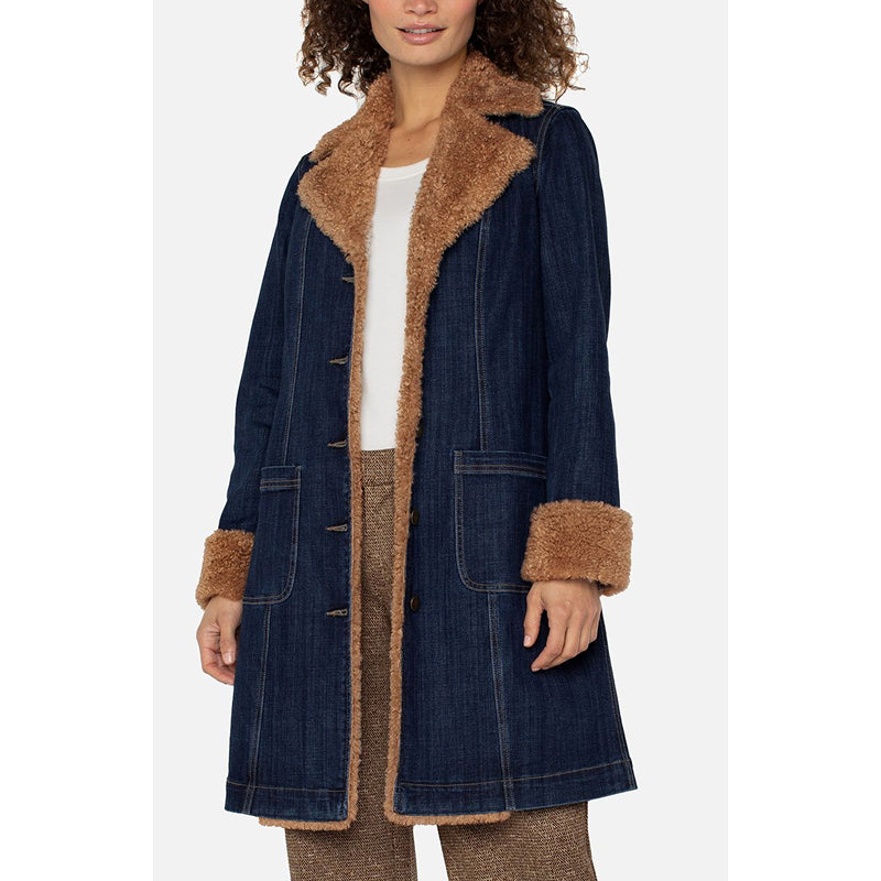 A woman wearing a dark blue denim-style coat with a tan faux-fur collar, front trim, and cuffs, over a white top and brown pants, photographed against a light gray studio background.