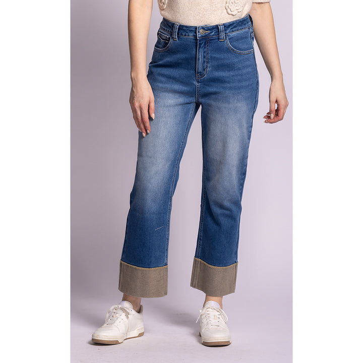A model wearing a pair of medium-wash blue cropped jeans with rolled cuffs showing a beige lining, paired with white sneakers and a cream top against a light lavender studio background.