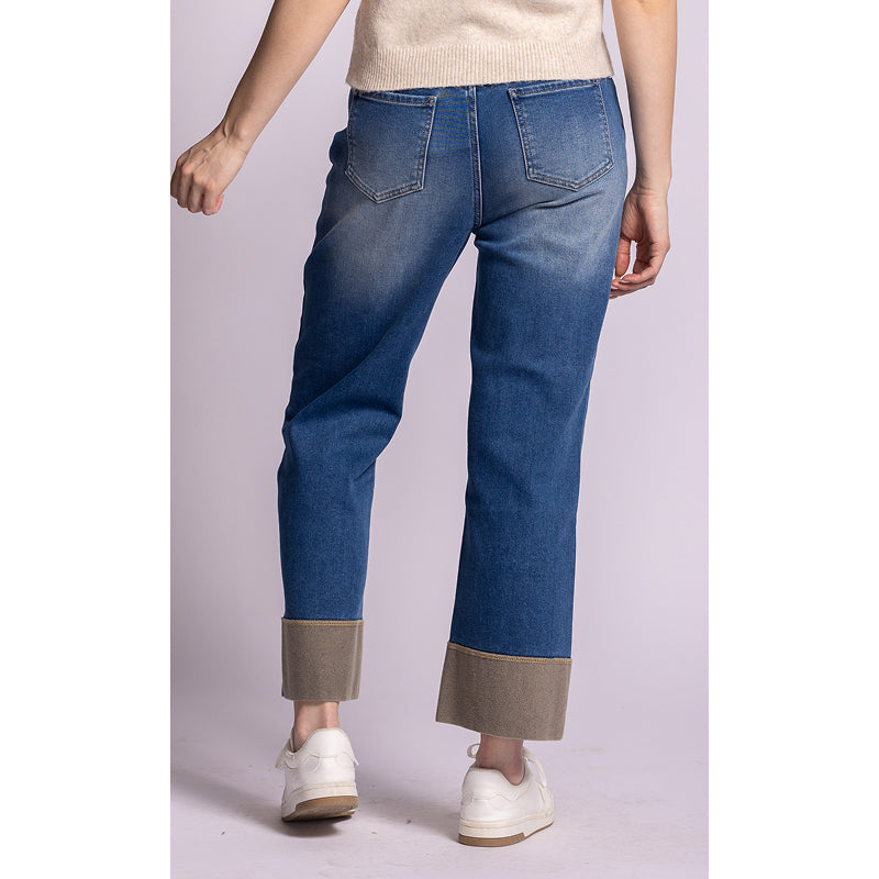 A person shown from the back wearing blue denim jeans with rolled cuffs and a contrasting beige lining, paired with a cream sweater and white sneakers against a pale lavender studio background.