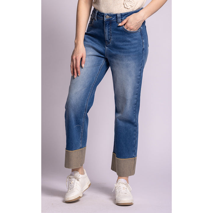 A model wearing a pair of medium-wash blue jeans with a unique contrast tan cuff, paired with white sneakers and a cream top, set against a solid light lavender background.