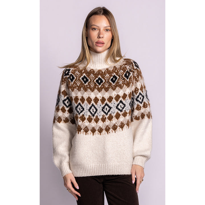 A woman modeling a cream-colored knit turtleneck sweater with a brown and black geometric Fair Isle pattern across the chest and shoulders, paired with dark brown pants, set against a solid light lavender background.
