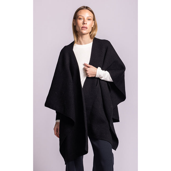 A woman modeling a black open-front poncho-style cardigan over a white long-sleeve top and dark pants, standing against a light lavender studio background.