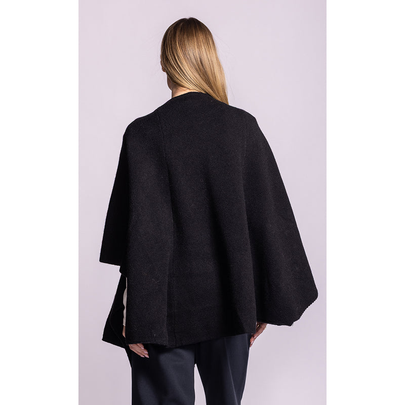 A woman shown from the back wearing a black open-front cape-style jacket with wide draped sleeves, paired with dark pants, against a light lavender studio background.