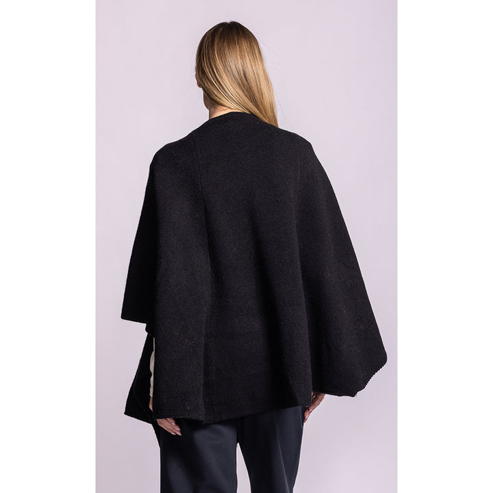 A woman shown from the back wearing a black open-front cape-style jacket with wide draped sleeves, paired with dark pants, against a light lavender studio background.