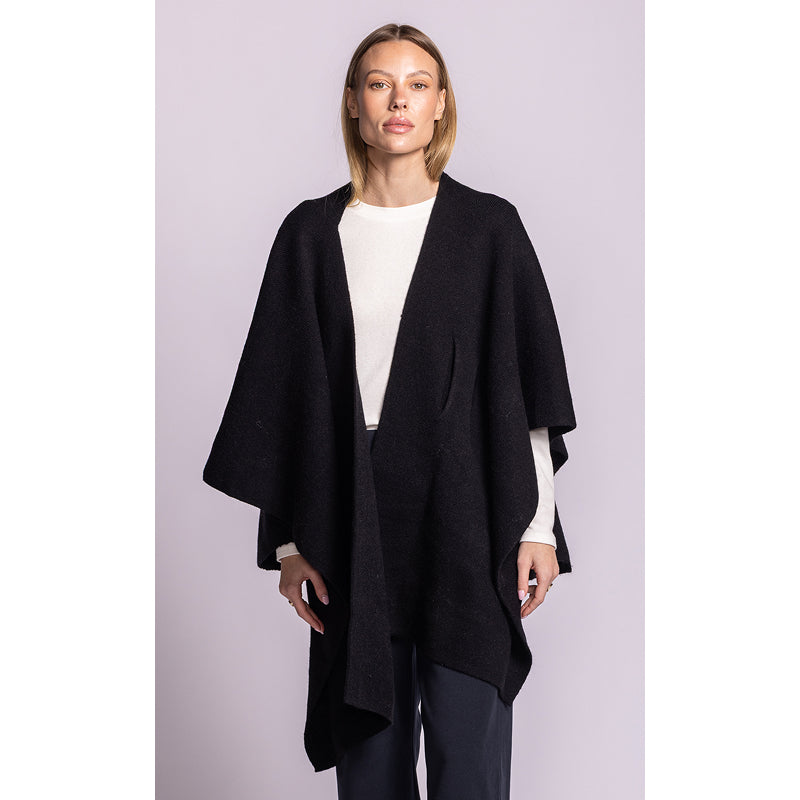 A woman modeling a black open-front poncho-style cardigan over a white top and dark trousers, standing against a light lavender studio background.