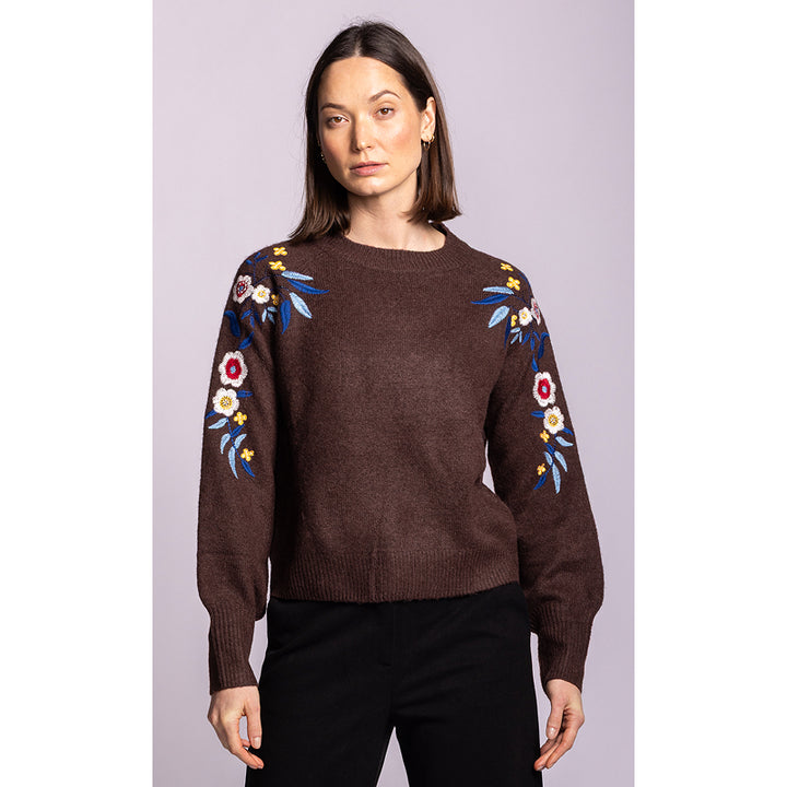A woman modeling a brown crewneck sweater with colorful floral embroidery on the upper sleeves, paired with black pants, against a solid light purple background.