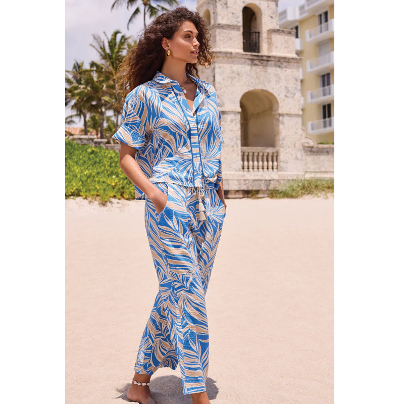 Woman in a blue and white patterned outfit standing on a sandy beach with palm trees and a building in the background.