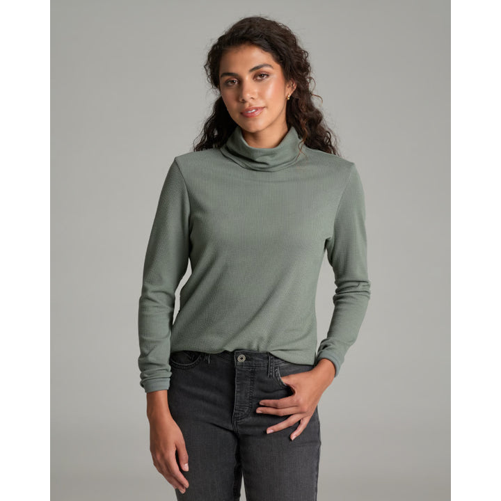 A woman modeling a sage green long-sleeve turtleneck top with dark gray jeans, standing against a solid gray studio background.