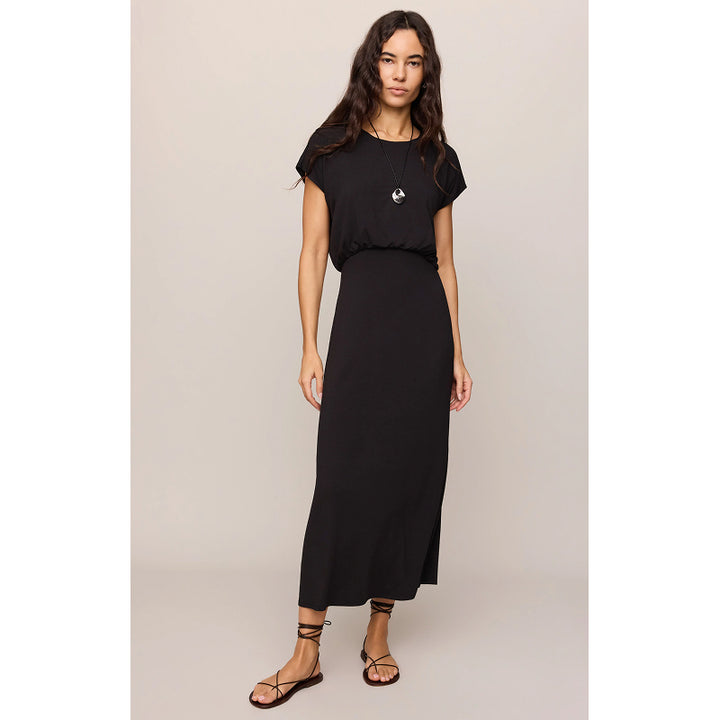 A woman modeling a black midi-length maxi dress with short sleeves and a small white circular logo on the chest, paired with black strappy flat sandals, against a solid light beige studio background.