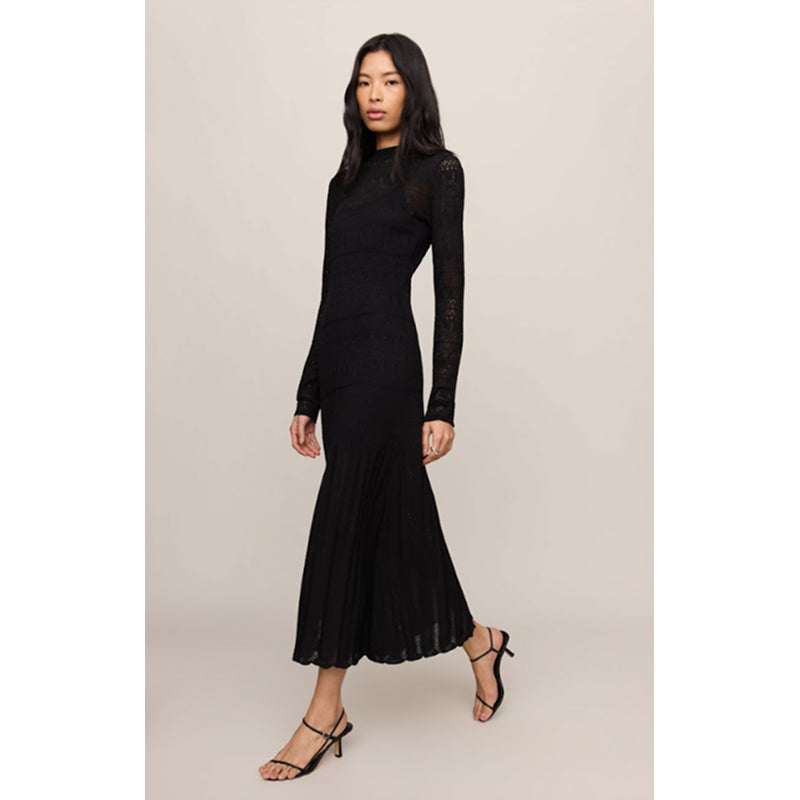 A woman modeling a black long-sleeve midi dress with sheer lace sleeves and a pleated skirt, paired with black strappy heels, against a solid light beige background.