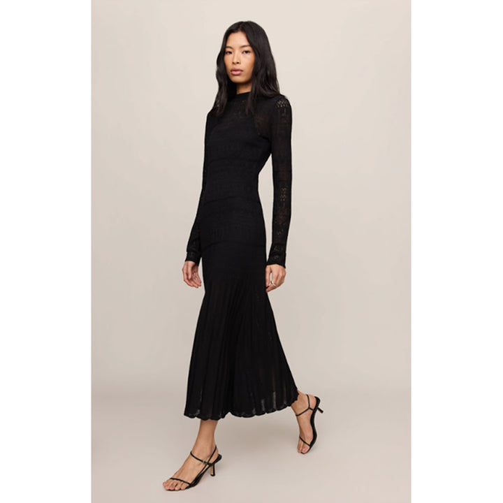 A woman modeling a black long-sleeve midi dress with sheer lace sleeves and a pleated skirt, paired with black strappy heels, against a solid light beige background.