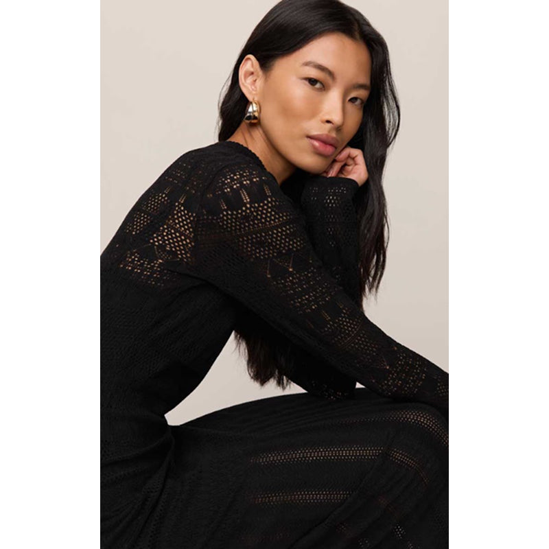 A woman wearing a black long-sleeve knit dress with an open-knit lace pattern, posed against a neutral beige background.