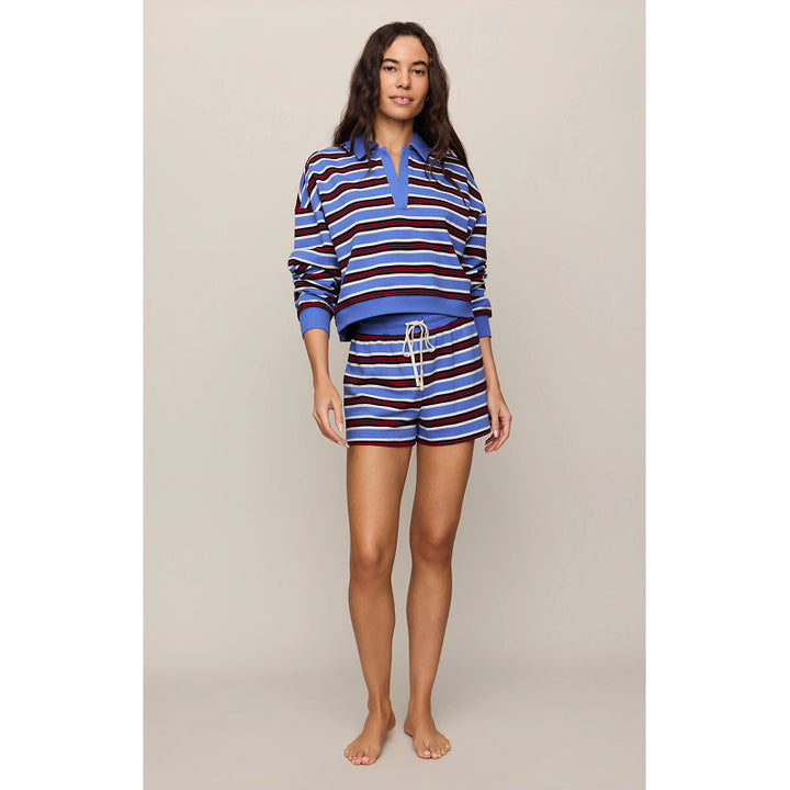 A woman modeling a matching two-piece lounge set featuring a long-sleeve rugby-style top and shorts, both with blue, navy, and white horizontal stripes, against a solid light beige studio background.