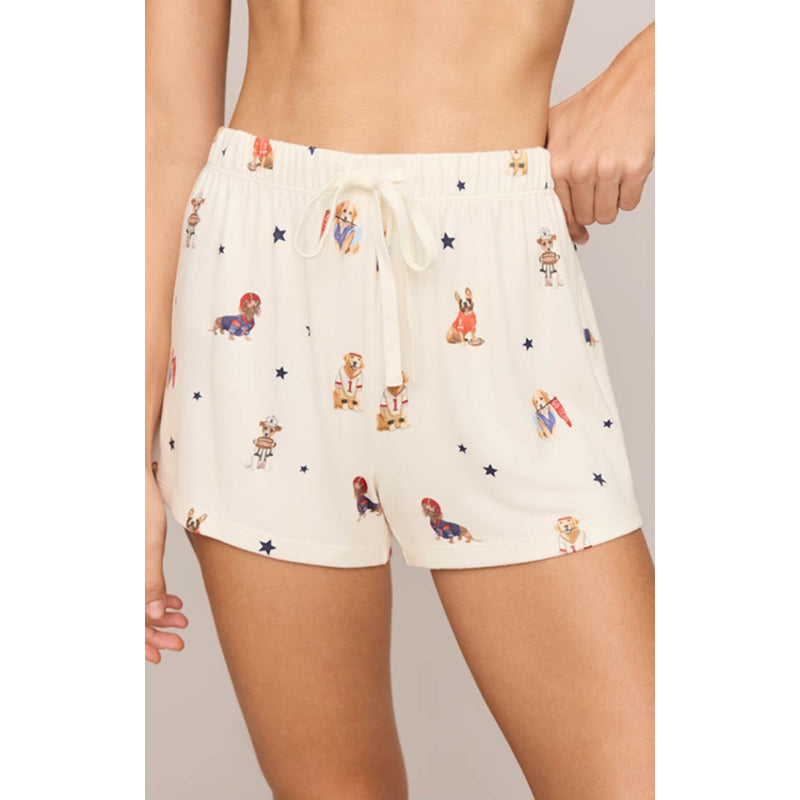 A person wearing cream-colored shorts with a whimsical print of cartoon dogs and small blue stars, shown from the waist to mid-thigh against a plain light gray studio background.
