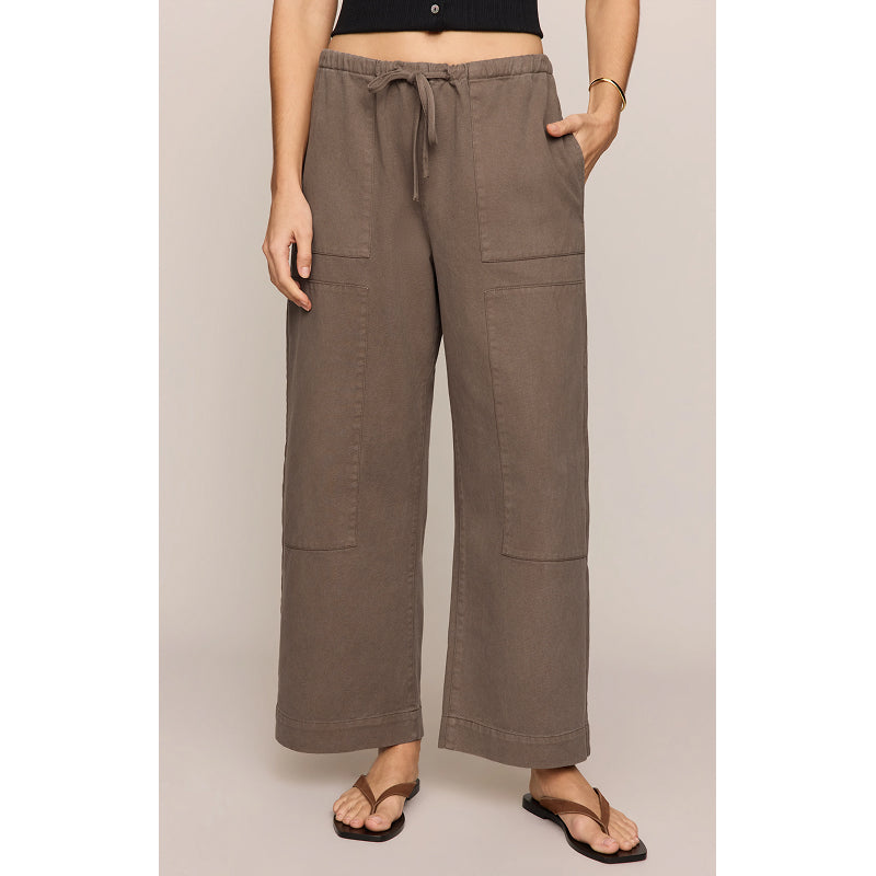 A model wearing wide-leg taupe pants with a drawstring waist, large front patch pockets, and a relaxed straight-leg fit, paired with brown flip-flops against a solid light beige background.