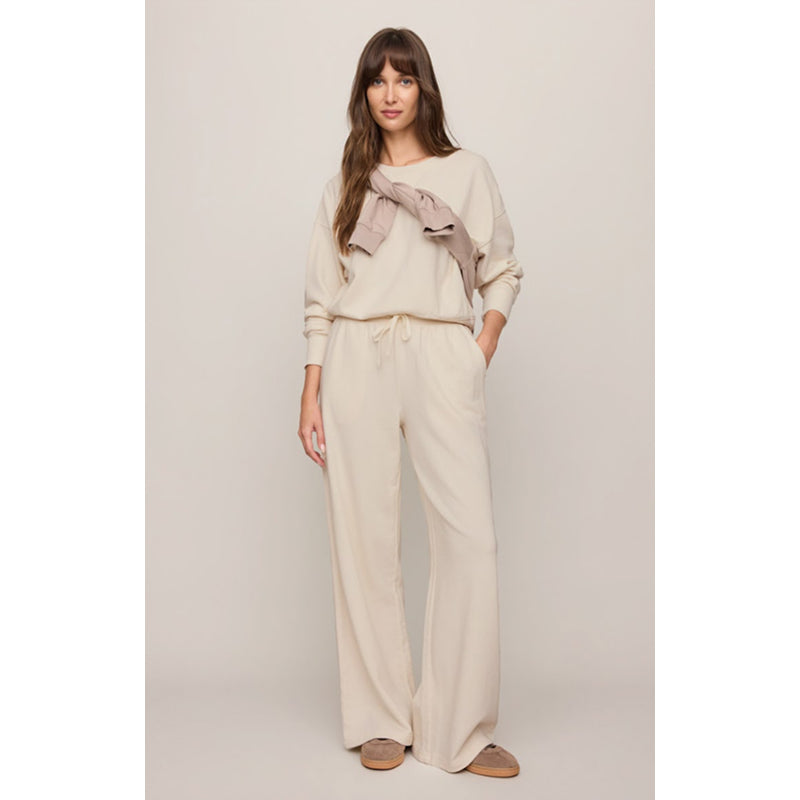 A woman modeling a matching beige lounge set consisting of a long-sleeve top  and matching wide-leg drawstring pants, paired with brown slip-on shoes against a solid light beige background.