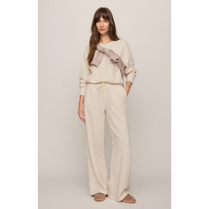 A woman modeling a matching beige lounge set consisting of a long-sleeve top  and matching wide-leg drawstring pants, paired with brown slip-on shoes against a solid light beige background.