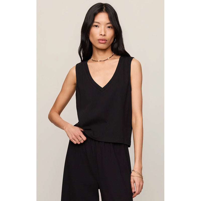 A woman modeling a black sleeveless V-neck top with a relaxed fit, paired with black pants, against a light beige studio background.
