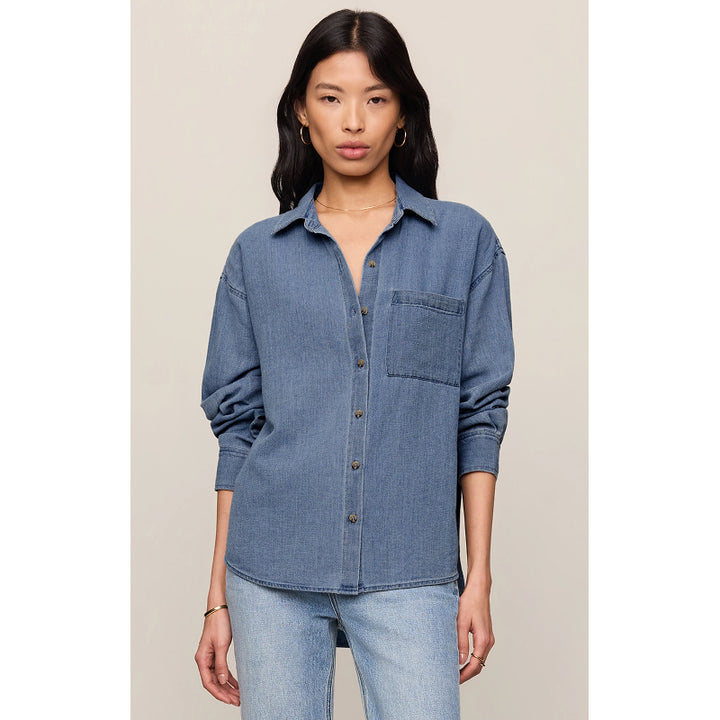 A woman modeling a medium-wash blue denim button-up shirt with a chest pocket, paired with light-wash jeans, against a solid light beige background.