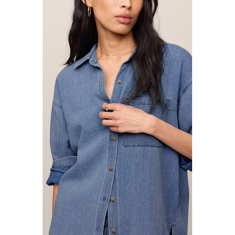 A woman wearing an oversized blue denim button-up shirt, shown from the chest down against a plain light beige background.
